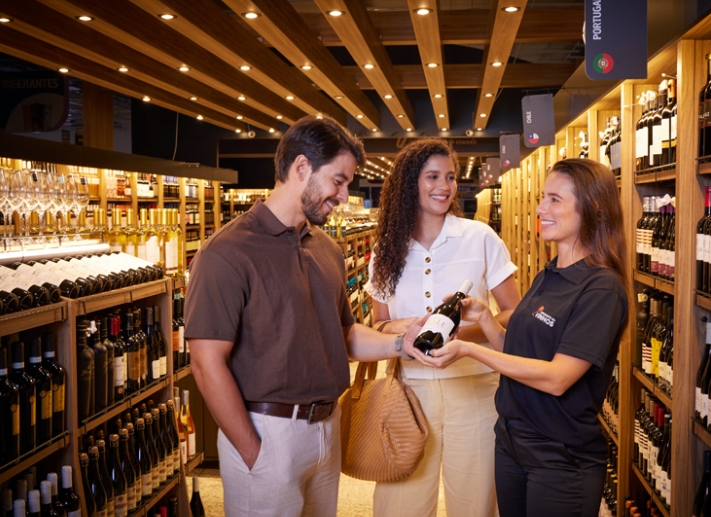 Especialista em vinhos do Guanabara apresentando uma garrafa de vinho tinto para um casal de clientes no corredor de vinhos da loja